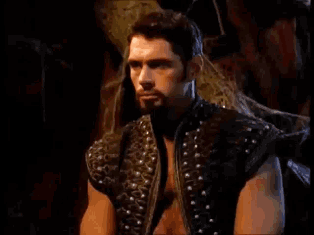 a man with a beard is sitting in a dark room wearing a black vest with studs .
