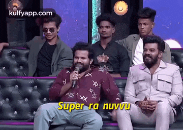 a group of men are sitting on a couch and one of them is holding a microphone and talking into it .