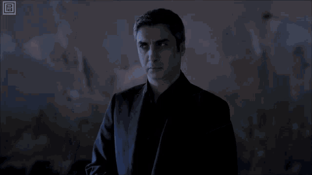 a man in a black suit stands in a dark room with a yellow rectangle in the corner