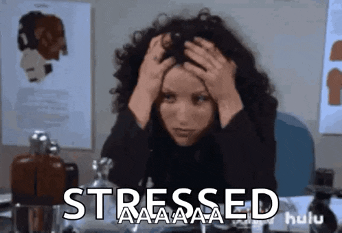 a woman with curly hair is sitting at a desk with her hands on her head and the words `` stressed '' written above her .