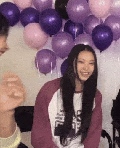 a woman wearing a pink and white shirt with the letter s on it is smiling in front of purple and pink balloons