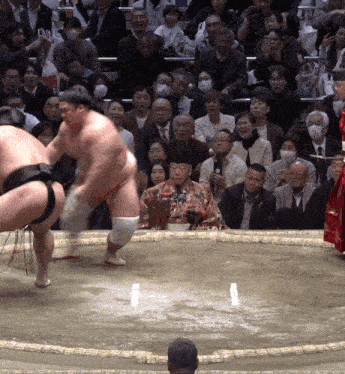 a sumo wrestler stands in front of a crowd while another wrestler looks on