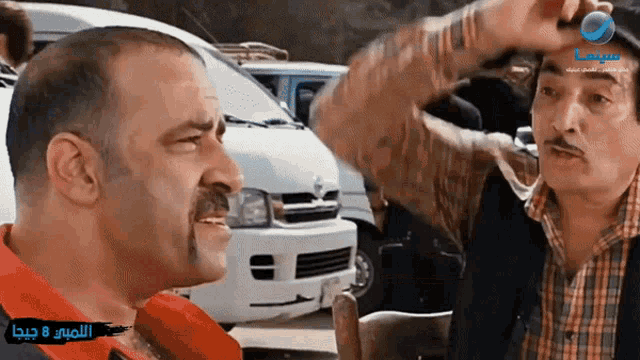 two men are talking in front of a white van with arabic writing on the bottom right corner