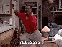 a man is dancing in a living room while wearing a red shirt and white pants .