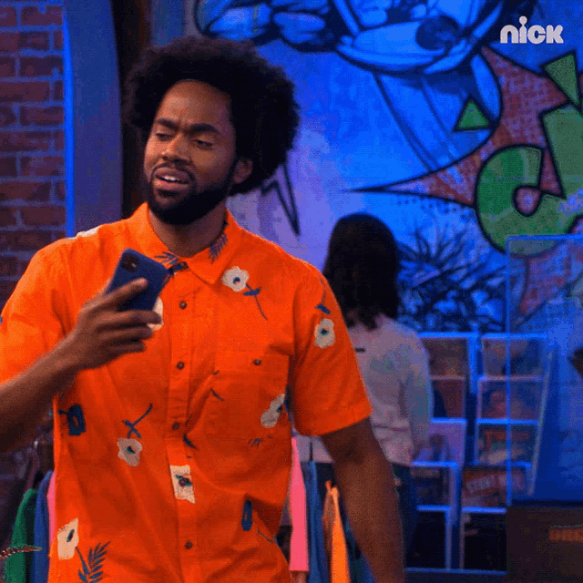 a man in an orange shirt is holding a cell phone in front of a brick wall with a nick logo on it