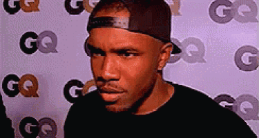 a man wearing a hat and a black shirt is standing in front of a wall with gq logos .