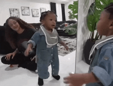 a baby is standing in front of a mirror in a living room while a woman sits on the floor .