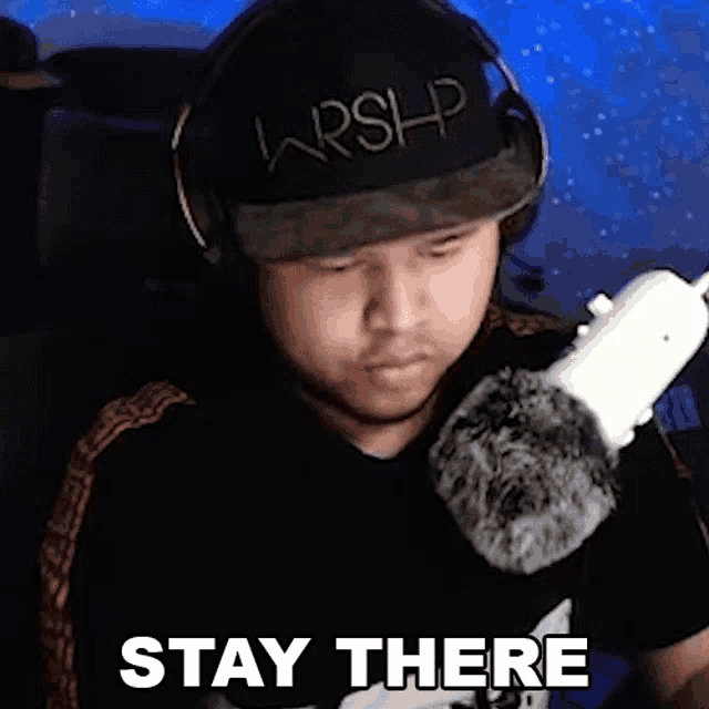 a man wearing headphones and a hat is standing in front of a microphone and says `` stay there '' .