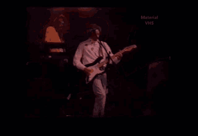 a man in a white shirt is playing a guitar in front of a microphone with the words material vhs above him