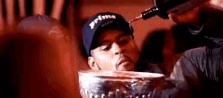 a man in a hat is drinking from a glass while another man pours liquid into it .