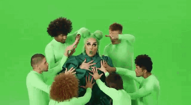 a drag queen is surrounded by a group of green dancers .