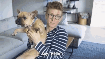 a man in a striped shirt is holding a dog and says " say hello "
