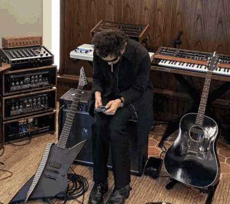a man in a black coat sits in front of a keyboard and guitar