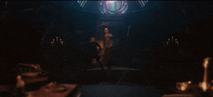 a man in a purple jacket is sitting at a table with his hands folded in a dark room .