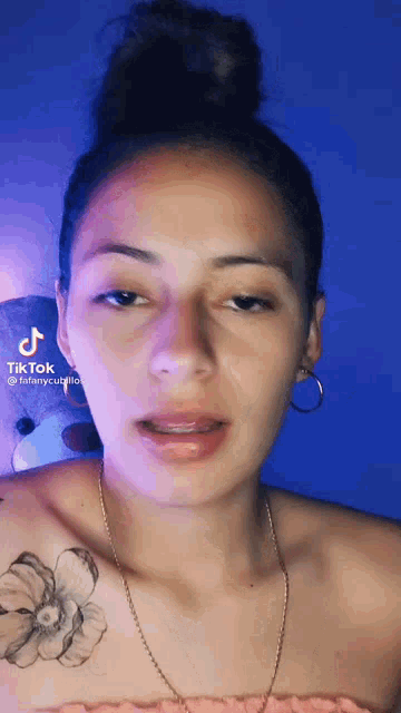 a woman with a bun on her head and a tattoo on her shoulder is talking to the camera .