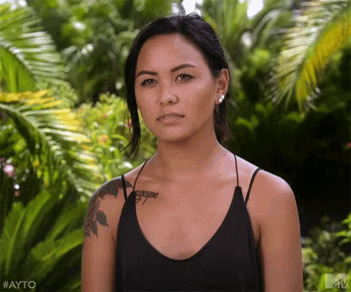 a woman with a tattoo on her shoulder is standing in front of a lush green forest .