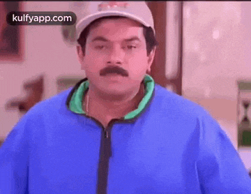 a man with a mustache wearing a baseball cap and a blue jacket is making a funny face .