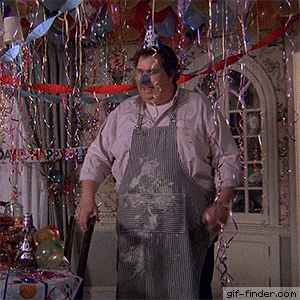 a man wearing an apron and a party hat is standing in front of a table with balloons and streamers