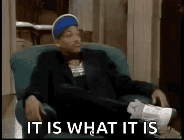 a man in a suit and blue hat is sitting in a chair with his feet up and the words `` it is what it is '' .