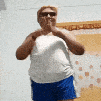 a man in a white tank top and blue shorts is standing in a room and making a funny face .