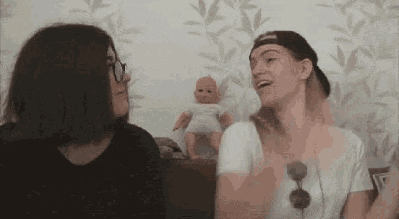 two women are sitting next to each other on a couch and talking to each other in front of a doll .