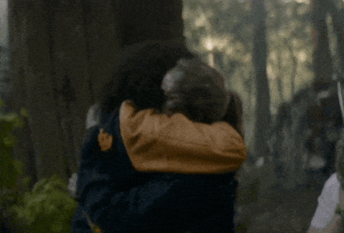 a woman in a blue jacket is being held by a man in a yellow jacket in the woods .
