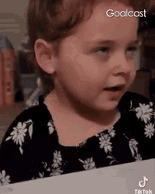 a little girl in a black and white floral dress is sitting at a table .