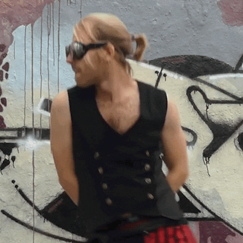 a man wearing sunglasses and a black vest stands in front of a graffiti wall