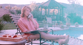 a woman in a pink shirt is laying on a lounge chair by a pool talking on a cell phone