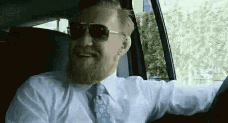 a man with a beard wearing sunglasses and a tie is driving a car .