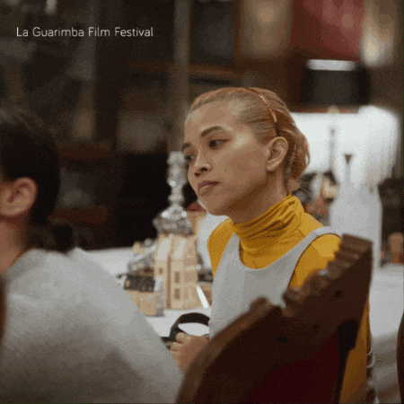 a woman in a yellow turtleneck sits at a table with a la guarimba film festival advertisement behind her