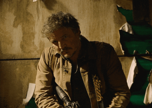a man in a tan jacket holds a gun in front of a green wall