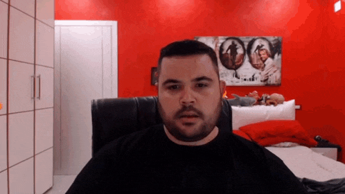 a man with a beard is sitting in front of a red wall with a picture on it