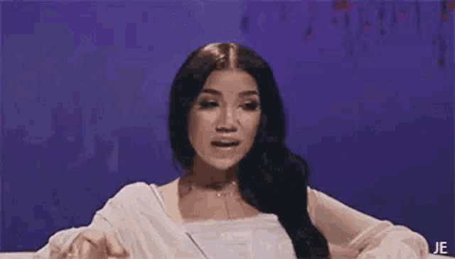 a woman with long black hair is sitting in front of a purple wall and making a funny face .
