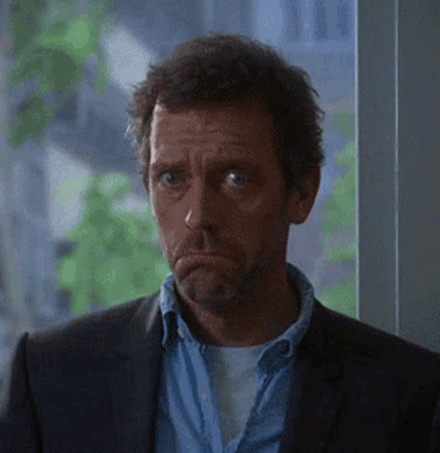 a man in a suit and blue shirt is standing in front of a window making a sad face .