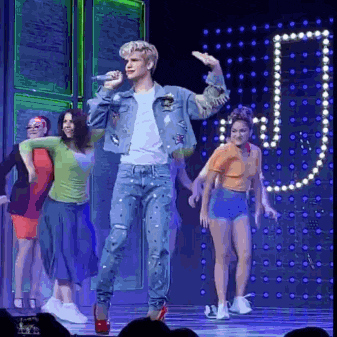 a man in a denim jacket is singing into a microphone on a stage surrounded by dancers