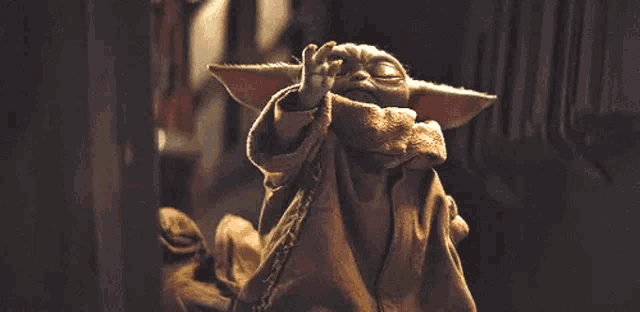 a close up of a baby yoda giving a peace sign while sitting down .
