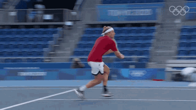 a man in a red shirt and white shorts is running on a tennis court with the olympic rings in the background