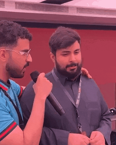 a man with a beard is talking into a microphone while another man holds his arm around his shoulder .