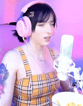 a woman wearing pink headphones and a plaid top sings into a white microphone