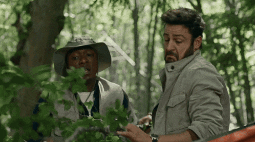 two men are standing in the woods one is wearing a hat and the other is wearing a watch