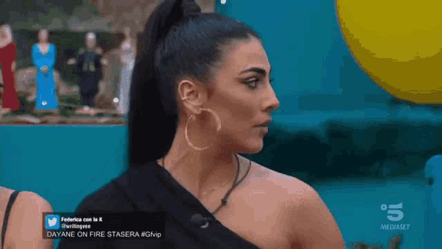 a woman wearing hoop earrings and a black one shoulder top is on a television screen that says 5 mediaset