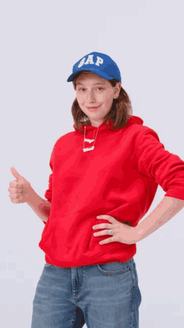 a woman wearing a red gap hoodie and a blue hat gives a thumbs up