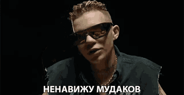 a man wearing sunglasses and a black vest is smoking a cigarette in a dark room .