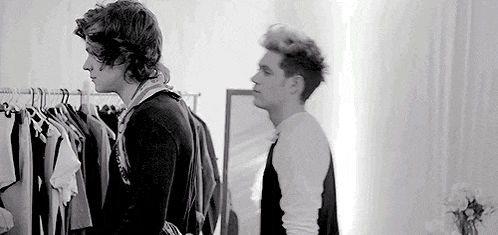 a black and white photo of two men standing next to each other in a room with clothes hanging on a rack .
