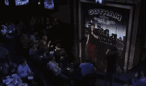 a man stands on a stage in front of a gotham comedy club sign