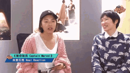 two women are sitting next to each other in front of a sign that says captain marvel