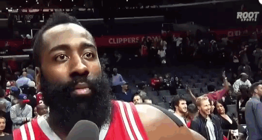 a man with a beard is talking into a microphone in front of a crowd at a basketball game
