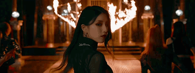 a woman in a black top stands in front of a fire in a dark room