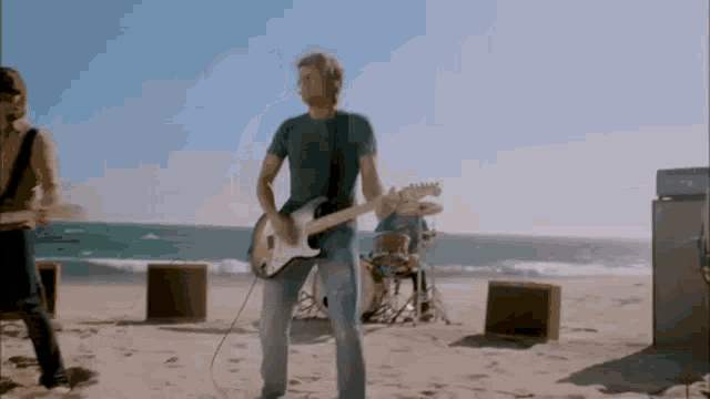 a man is playing a guitar on the beach while a band plays drums .